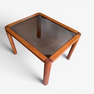 May include: A rectangular side table featuring a warm-toned wooden frame with rounded corners and four cylindrical legs. The tabletop consists of a dark, semi-transparent smoked glass panel set flush within the wooden border structure.
