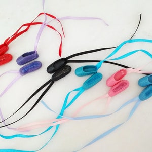 Puede incluir: Una colección de ocho zapatillas de ballet en miniatura con cintas de varios colores. Las zapatillas están hechas de plástico y tienen un acabado brillante. Las cintas están atadas en lazos.