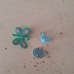 Może przedstawiać: Kolekcja zabawek Polly Pocket w stylu vintage. Zawiera zielonego motyla z różowymi akcentami, jasnoniebieskiego łabędzia i niebieską owalną zawieszkę z napisem "Polly Pocket".