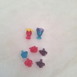以下が含まれることがあります： 小さくてカラフルな半透明のプラスチック製おもちゃのコレクション。2つはサンデーグラスの形で、1つは赤いトッピング、もう1つは紫色のトッピング。他の6つは花形で、赤、青、紫の色合いです。