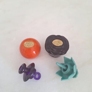 May include: Four decorative bottle stoppers. Two are round, one orange and one dark brown, each with a gold emblem. The other two are purple and teal, with unique shapes. These stoppers are designed for wine bottles.