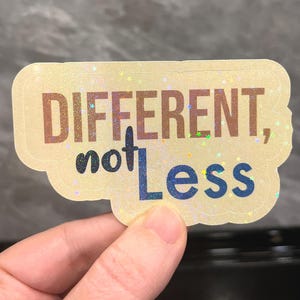 Puede incluir: Una pegatina holográfica con las palabras "DIFFERENT, not Less" en letras marrones y azules. La pegatina tiene un fondo amarillo claro y un borde decorativo. La pegatina está sujeta por una mano.