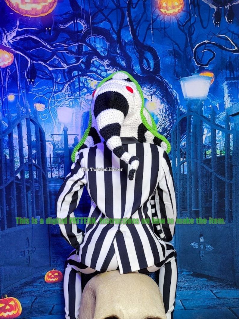 May include: A black and white striped suit with a green and white crocheted hat. The hat is shaped like a long, pointed cone with a red pom-pom on top. The suit is sitting on a white skull. The background is a blue and green Halloween-themed backdrop with pumpkins and a gate.