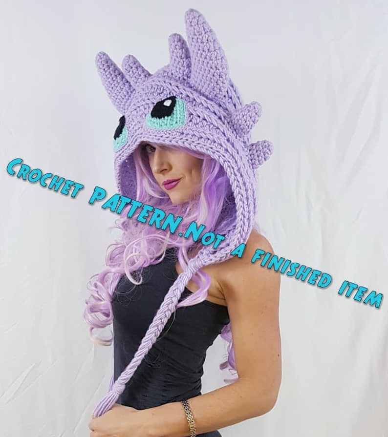 K&ouml;nnte beinhalten: H&auml;kelanleitung f&uuml;r eine lilafarbene Drachen-Kapuzenm&uuml;tze mit einem langen Zopf. Das Muster ist kein fertiges Produkt. "Crochet Pattern. Not a Finished Item" steht in blauer Schrift auf dem Bild.