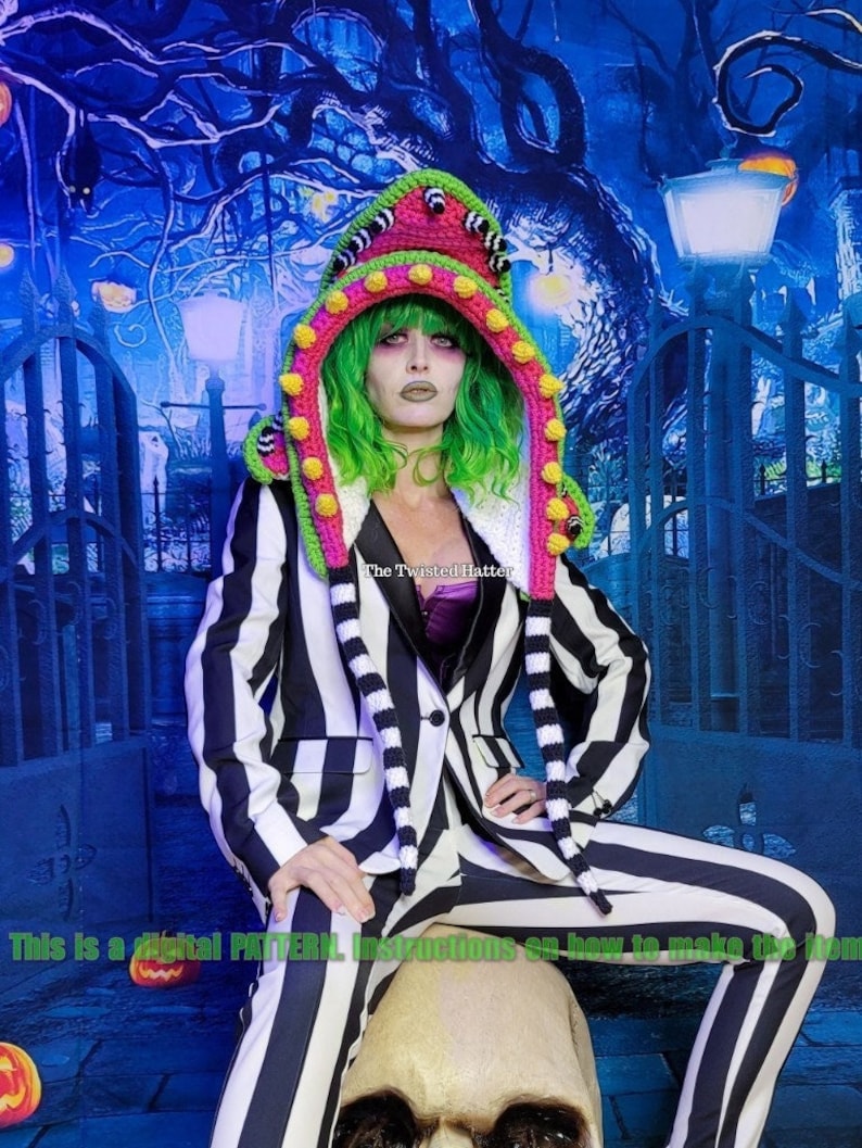 May include: A crochet hat with a green and pink striped design and a large, white pom-pom on top. The hat is shaped like a monster with large, black eyes and a pink mouth. The hat is worn by a person with green hair and a black and white striped outfit. The text "The Twisted Hatter" is visible on the hat.