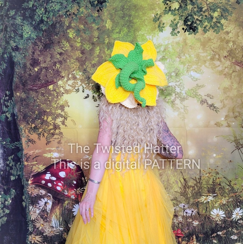May include: A crocheted yellow flower hat with a green dragon design. The hat is worn by a person in a yellow dress. The background is a blurred image of a forest.