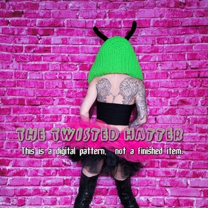 May include: A person wearing a green crocheted hat with horns, a black top, and a pink tulle skirt. The text "THE TWISTED HATTER" and "This is a digital pattern, not a finished item." is displayed below the person.