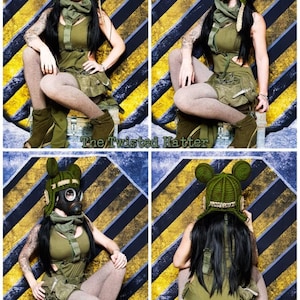May include: A woman wearing a green and brown outfit with a gas mask and a crocheted hat with ears. The hat has the word "Wonderland" written on it. She is sitting on a box in front of a yellow and black striped background.