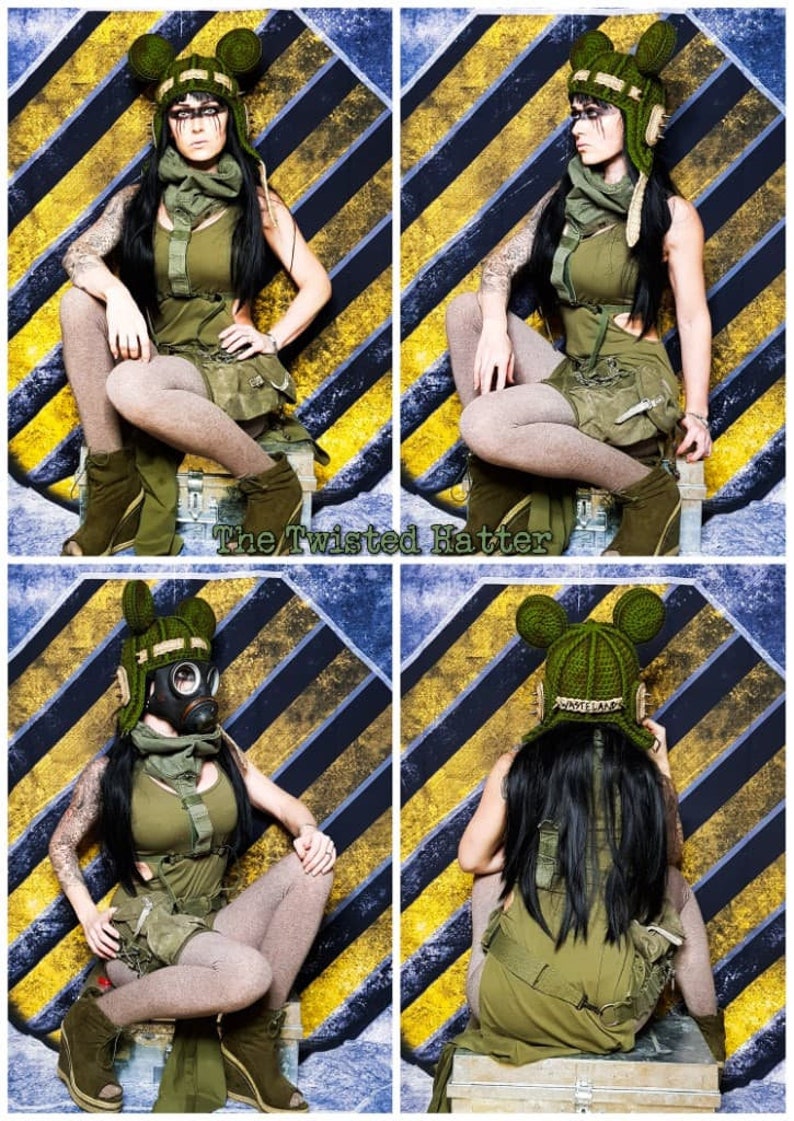 May include: A woman wearing a green and brown outfit with a gas mask and a crocheted hat with ears. The hat has the word "Wonderland" written on it. She is sitting on a box in front of a yellow and black striped background.