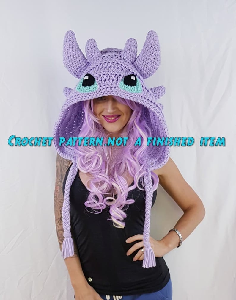 K&ouml;nnte beinhalten: H&auml;kelanleitung f&uuml;r eine lilafarbene Drachen-Kapuzenm&uuml;tze mit t&uuml;rkisfarbenen Augen. Das Muster ist kein fertiges Produkt.  "CROCHET PATTERN. NOT A FINISHED ITEM" steht auf dem Bild.