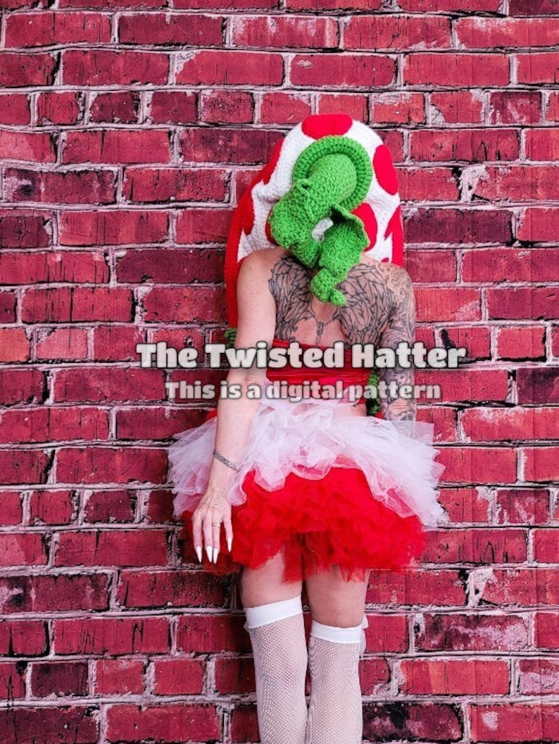May include: A person wearing a red and white tutu skirt with a green and white crocheted mushroom hat. The background is a red brick wall. The text "The Twisted Hatter" and "This is a digital pattern" is visible on the image.