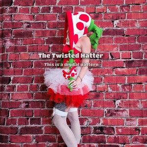 May include: A woman wearing a red and white crocheted mushroom hat with green leaves, a white tulle skirt with red trim, and white thigh-high stockings. The background is a red brick wall. The text "The Twisted Hatter" and "This is a digital pattern" is visible in the image.