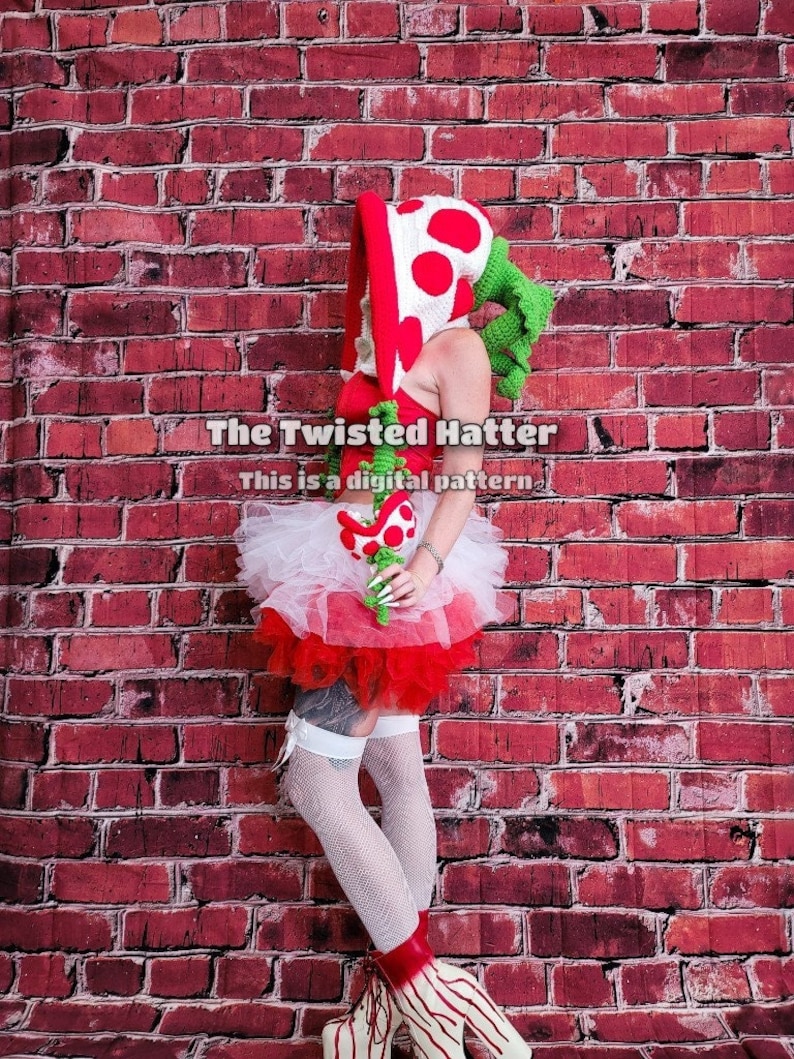 May include: A woman wearing a red and white crocheted mushroom hat with green leaves, a white tulle skirt with red trim, and white thigh-high stockings. The background is a red brick wall. The text "The Twisted Hatter" and "This is a digital pattern" is visible in the image.