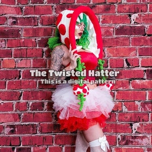 May include: A woman wearing a crocheted green and red plant costume with a white tutu and white stockings. The costume is inspired by a video game character. The text "The Twisted Hatter This is a digital pattern" is visible in the image.