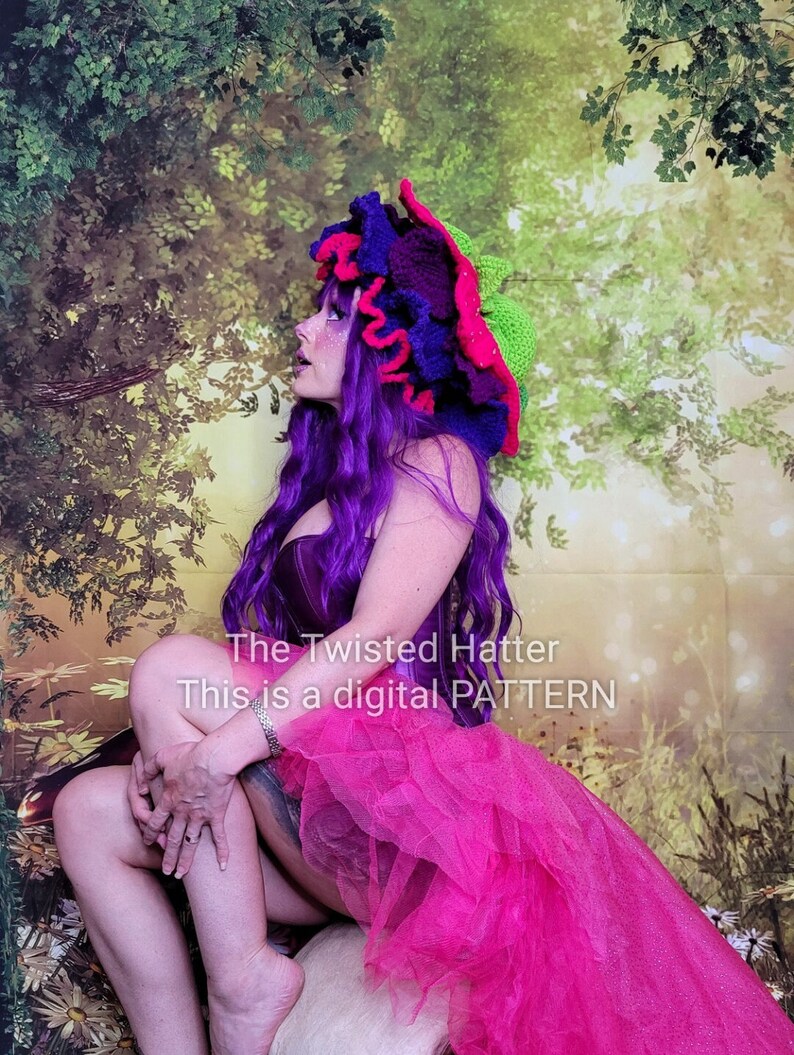May include: A woman wearing a bright pink and purple crocheted hat with a green brim. The hat is shaped like a mushroom. The woman is wearing a purple corset and a pink tulle skirt. The background is a green and brown leafy pattern.