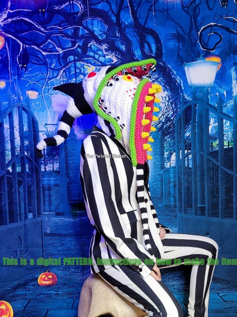 May include: A crochet pattern for a colourful, whimsical hat featuring a large, toothy mouth with eyes and a long, striped tail. The hat is worn by a person in a black and white striped suit, sitting on a white, skull-shaped stool. The background is a blue and green forest scene with a gate and a street lamp.