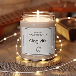 May include: A brown candle in a glass jar with a white label that reads "The not so calming scent of Gingivitis" with a tooth graphic. The candle is lit and sitting on a wooden coaster.