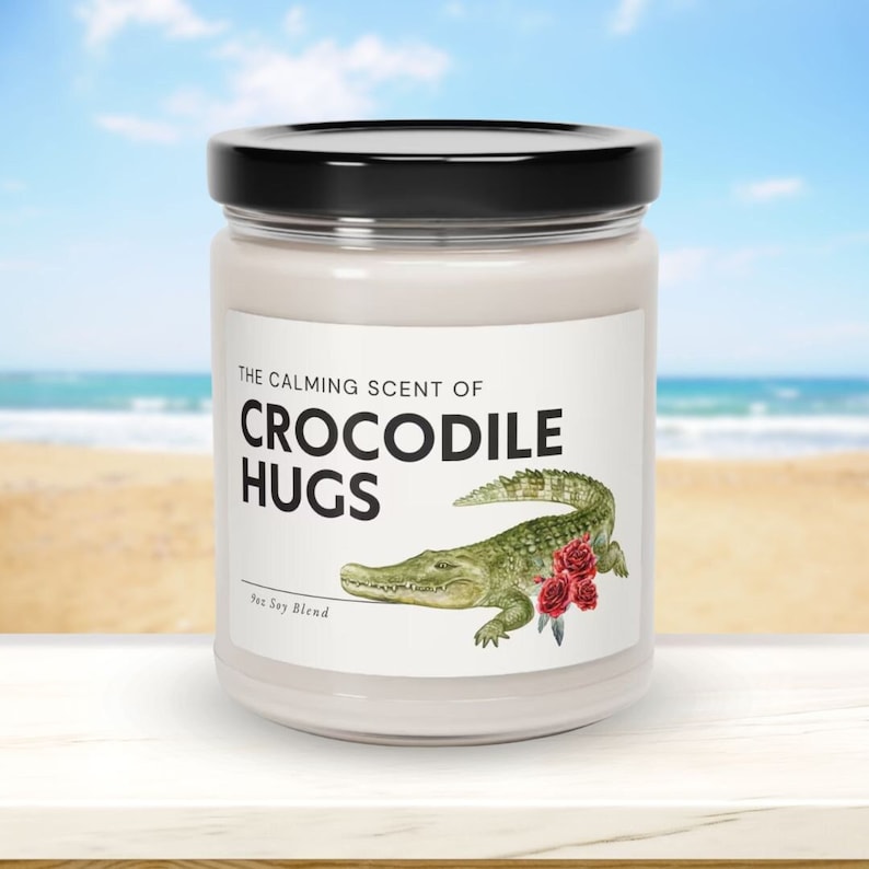May include: A white candle jar with a black lid. The label reads "The Calming Scent of Crocodile Hugs" with a green crocodile illustration and red roses. The candle is a soy blend.