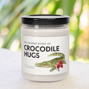 May include: A white candle jar with a black lid. The label reads "The Calming Scent of Crocodile Hugs" with a green crocodile illustration and red roses. The label also says "9oz Soy Blend".