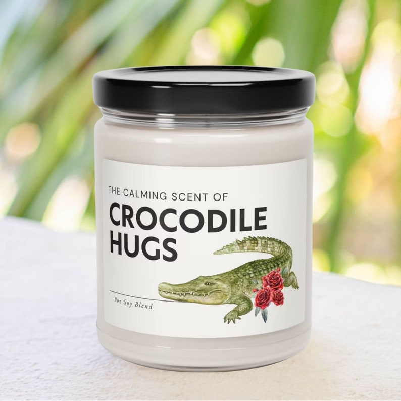 May include: A white candle jar with a black lid. The label reads "The Calming Scent of Crocodile Hugs" with a green crocodile illustration and red roses. The label also says "9oz Soy Blend".