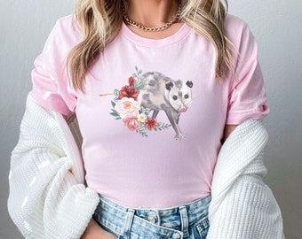 Pode incluir: Uma camiseta rosa com um design floral e um gráfico de gambá. O gambá é cinza e branco com o nariz e a língua rosa. As flores são rosa, vermelhas e brancas.