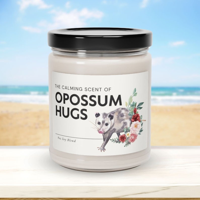 May include: A white candle jar with a black lid. The label reads "The Calming Scent of Opossum Hugs" with a watercolor illustration of an opossum and flowers. The text "100% Soy Blend" is also on the label.