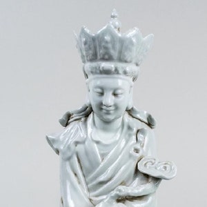 Puede incluir: Una serena estatua de cer&aacute;mica de color claro de una figura sentada, adornada con una corona y t&uacute;nicas fluidas. La figura est&aacute; sentada sobre una base en forma de nube, con las manos suavemente posadas. La estatua mide unos 25 cm de altura.