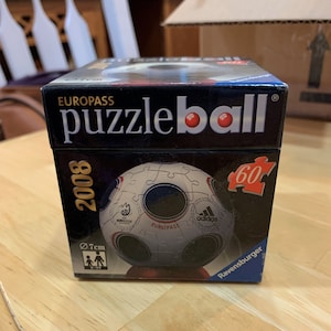 Puede incluir: Una caja negra con las palabras "EUROPASS puzzleball" impresas en la parte delantera. La caja presenta un rompecabezas de 60 piezas de un bal&oacute;n de f&uacute;tbol con el logotipo de Adidas y las palabras "EURO 2008" impresas en &eacute;l. La caja tiene un di&aacute;metro de 7 cm.