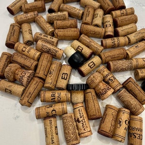 May include: A collection of used wine corks, some with labels and some without. The corks are various shades of brown and beige, and some have black or white markings. The corks are scattered on a white surface.