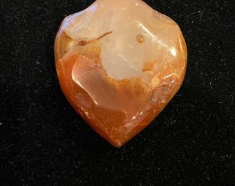 Puede incluir: Un colgante de cornalina en forma de corazón con una anilla de plata. La cornalina es de color naranja intenso con remolinos blancos y marrones.