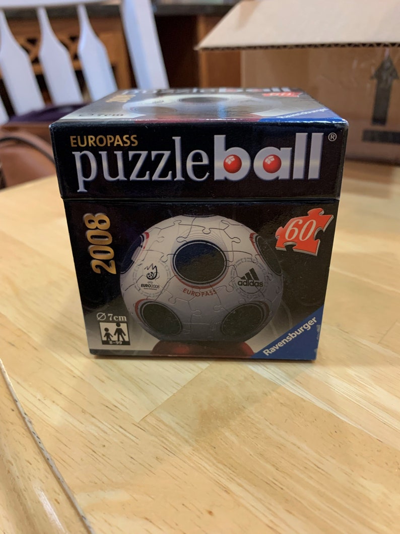Puede incluir: Una caja negra con las palabras "EUROPASS puzzleball" impresas en la parte delantera. La caja presenta un rompecabezas de 60 piezas de un bal&oacute;n de f&uacute;tbol con el logotipo de Adidas y las palabras "EURO 2008" impresas en &eacute;l. La caja tiene un di&aacute;metro de 7 cm.