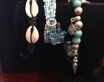 May include: Three bracelets with different styles. The first bracelet is made of white seashells and black cord. The second bracelet is made of blue and white beads. The third bracelet is made of turquoise beads and silver charms.