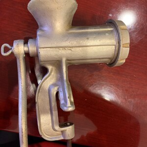 May include: A vintage, silver-colored meat grinder with a clamp base and a hand crank. The grinder has a funnel-shaped top, a cylindrical body, and a screw-on grinding plate. The handle is made of wood. The grinder is designed to be attached to a table.