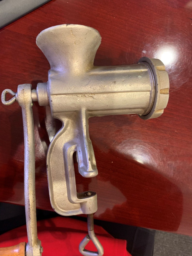 May include: A vintage, silver-colored meat grinder with a clamp base and a hand crank. The grinder has a funnel-shaped top, a cylindrical body, and a screw-on grinding plate. The handle is made of wood. The grinder is designed to be attached to a table.