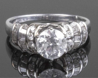 Könnte beinhalten: Ein silberner Ring mit einem großen, runden, klaren Diamanten in der Mitte, umgeben von kleineren Diamanten. Die Ringschiene ist mit Baguette-förmigen Diamanten besetzt.