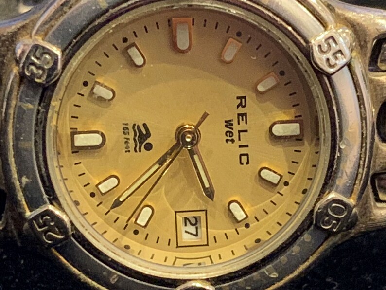 Peut inclure: Une montre-bracelet dor&eacute;e avec un cadran de couleur cr&egrave;me. Le cadran de la montre comporte des marques noires et des index blancs. Le cadran de la montre comporte &eacute;galement une fen&ecirc;tre de date &agrave; 3 heures et les mots "RELIC wet" imprim&eacute;s sur le cadran. La montre est &eacute;tanche &agrave; 165 pieds.