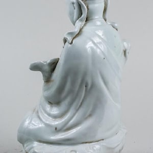 Puede incluir: Figura de cer&aacute;mica azul claro de una figura sentada, posiblemente una deidad, con tocado adornado y t&uacute;nicas fluidas. La figura est&aacute; sentada sobre una base texturizada. La figura mide aproximadamente 15 cm de altura.