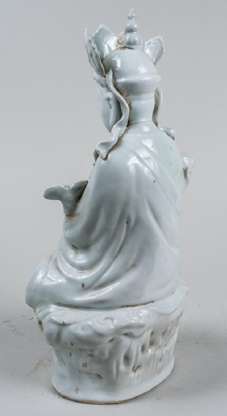 Puede incluir: Figura de cer&aacute;mica azul claro de una figura sentada, posiblemente una deidad, con tocado adornado y t&uacute;nicas fluidas. La figura est&aacute; sentada sobre una base texturizada. La figura mide aproximadamente 15 cm de altura.