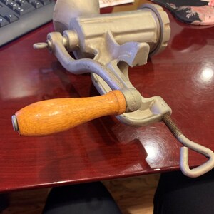 May include: A vintage, silver-colored meat grinder with a wooden handle. The grinder has a hand crank and a clamp for securing it to a surface. The handle is a light brown color. The grinder is made of metal and appears to be well-used.