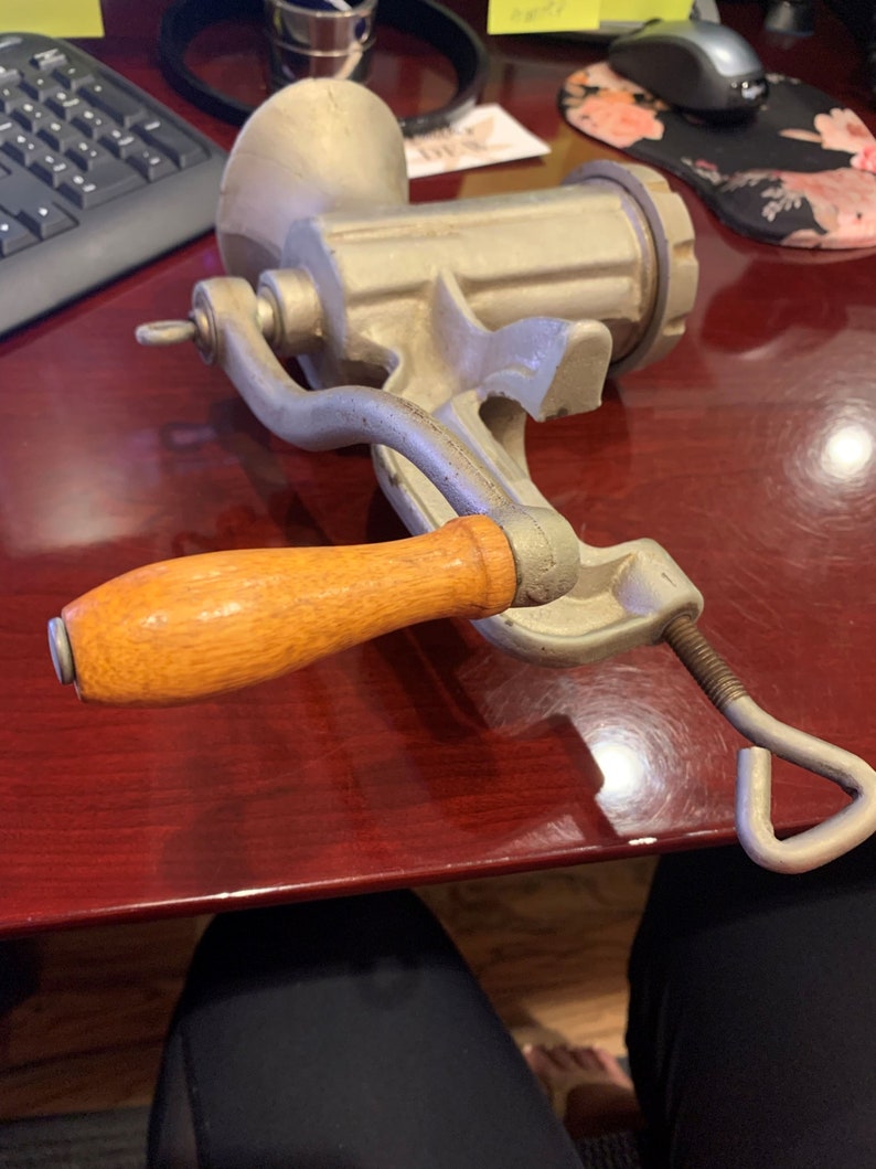 May include: A vintage, silver-colored meat grinder with a wooden handle. The grinder has a hand crank and a clamp for securing it to a surface. The handle is a light brown color. The grinder is made of metal and appears to be well-used.