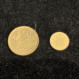 Puede incluir: Dos monedas de color dorado. La moneda m&aacute;s grande presenta un gallo y el texto "50 FRANCS - 1951 - LIBERTE EGALITE FRATERNITE". La moneda m&aacute;s peque&ntilde;a presenta un dise&ntilde;o de un &aacute;rbol y el texto "G".