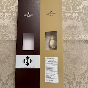 May include: Two brown and gold boxes with the Patek Philippe Geneve logo. The box on the right has a watch inside and a list of items included in the box. The list includes: 1 watch, 1 leather strap with buckle, 1 leather strap with clasp, 1 metal bracelet, 1 complete strap, 2 extra straps, 1 watch tool, 1 watch case back, and a visa operator.