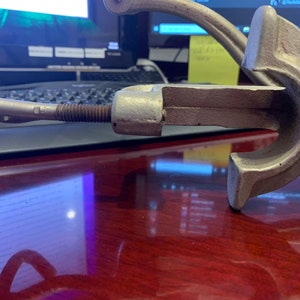 May include: A vintage, cast-iron clamp with a screw mechanism. The clamp is silver-colored with a handle and a threaded rod. The design suggests it is for woodworking or metalworking. The clamp is resting on a dark wooden surface.