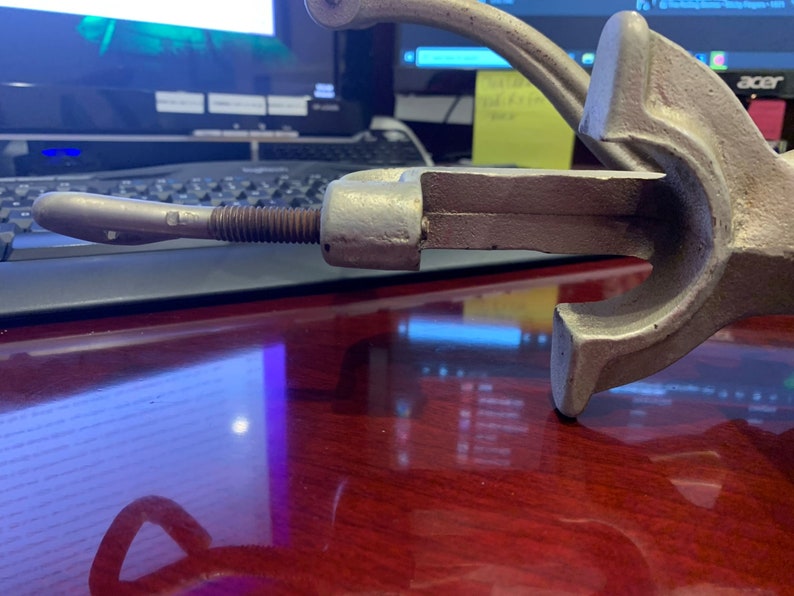 May include: A vintage, cast-iron clamp with a screw mechanism. The clamp is silver-colored with a handle and a threaded rod. The design suggests it is for woodworking or metalworking. The clamp is resting on a dark wooden surface.