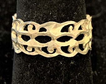 Puede incluir: Un anillo plateado con un diseño calado. El anillo presenta un patrón repetitivo de formas curvas y aberturas ovaladas. El anillo se muestra sobre una superficie negra, resaltando sus intrincados detalles. Una pieza de joyería decorativa.