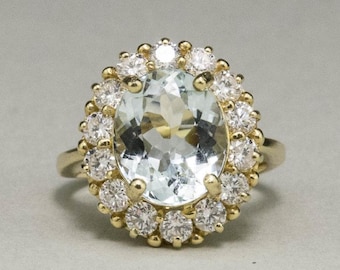 May include: A gold ring featuring a large, oval-cut, pale blue gemstone surrounded by a halo of smaller, round, clear gemstones. The ring's band is a simple, polished gold. The gemstones sparkle against a neutral background.