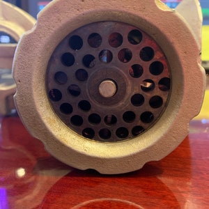 May include: Close-up of a vintage, off-white meat grinder. The grinder's head features a circular plate with numerous small holes, surrounded by a textured, ridged rim. The central grinding mechanism is visible, with a silver-colored screw and a central pin.