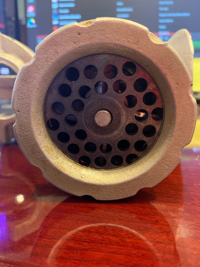 May include: Close-up of a vintage, off-white meat grinder. The grinder's head features a circular plate with numerous small holes, surrounded by a textured, ridged rim. The central grinding mechanism is visible, with a silver-colored screw and a central pin.