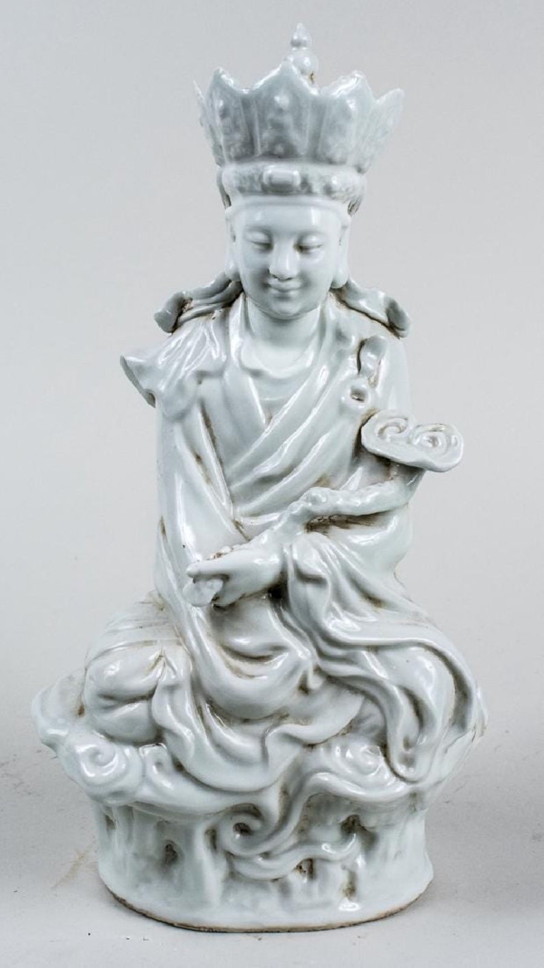 Puede incluir: Una serena estatua de cer&aacute;mica de color claro de una figura sentada, adornada con una corona y t&uacute;nicas fluidas. La figura est&aacute; sentada sobre una base en forma de nube, con las manos suavemente posadas. La estatua mide unos 25 cm de altura.