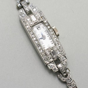 May include: A vintage rectangular wristwatch with a white face and silver hands. The watch is encrusted with numerous small diamonds, and the band is also adorned with diamonds. The watch has a classic, elegant design.